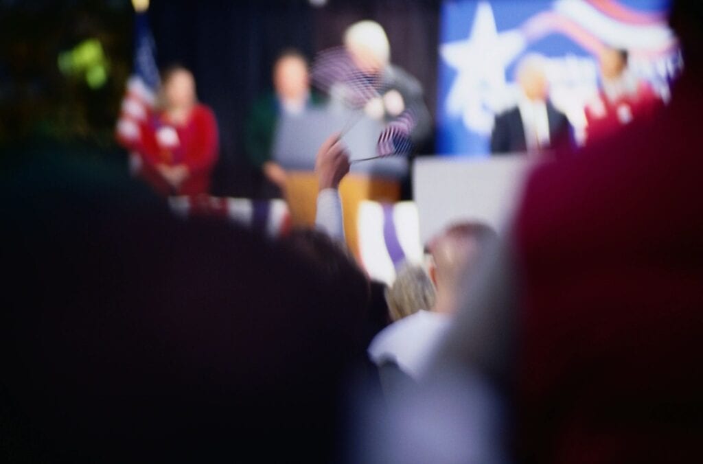 Blurred image of a person speaking at an event with audience in foreground.