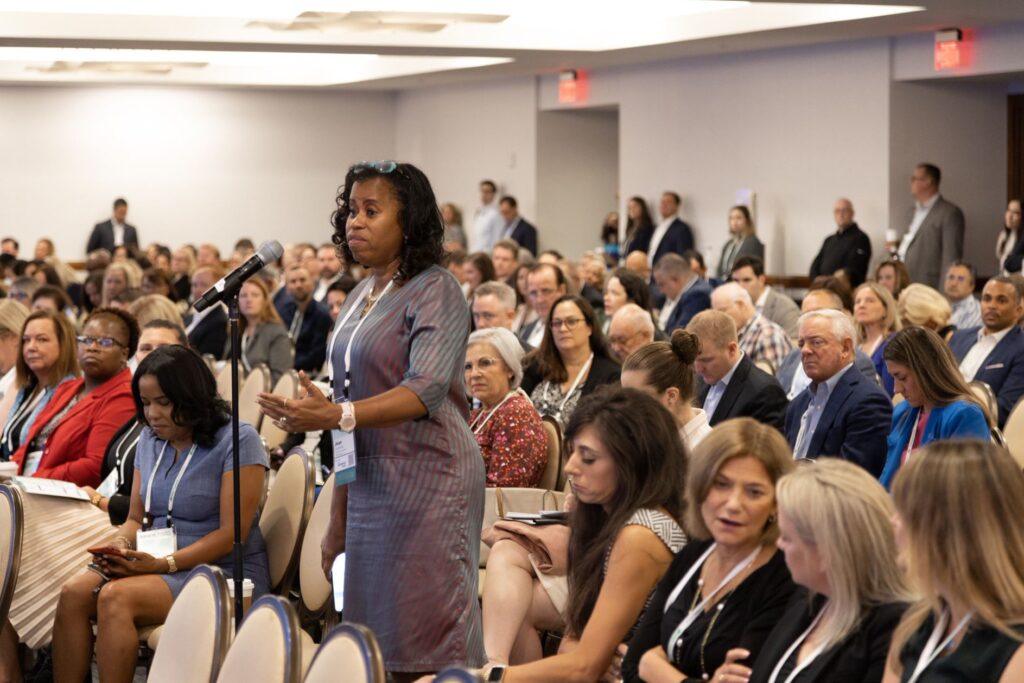 A woman stands speaking at a crowded conference or meeting.
