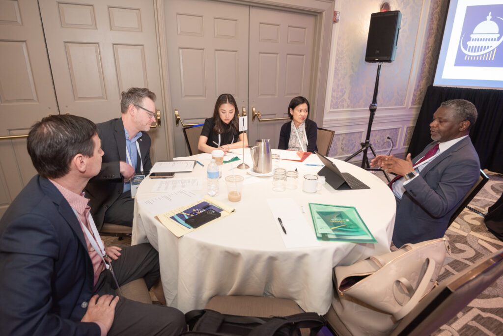 People engaged in a formal discussion around a round table in a conference room.